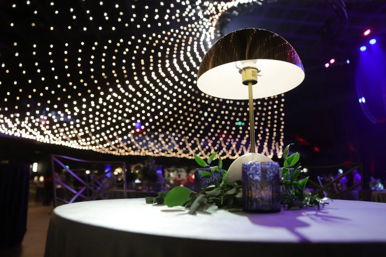 Gold dome cordless LED table lamp glowing beside eucalyptus sprigs and cobalt glass candle holders on white linen, fairy-light canopy blurred behind