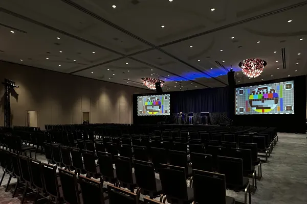 Hotel ballroom set theater-style for a general session, two projection screens and flown line-array speakers framing a draped stage