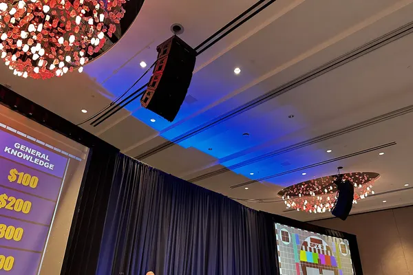 Line-array speaker hang flown over a ballroom stage with blue ceiling light and a game-show board on the side screen